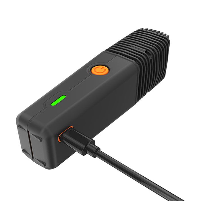 Angled close-up of a black portable vaporizer with a green indicator light and an orange power button, connected to a black charging cable.