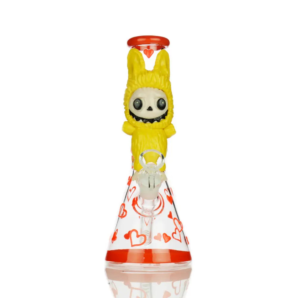 Front view of a clear glass beaker water pipe featuring red heart patterns, a red base, and a yellow character top resembling a skeleton in a bunny suit.