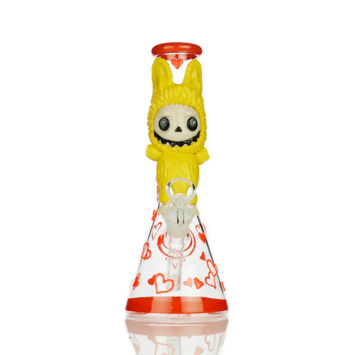 A 10-inch glass beaker water pipe featuring a yellow glow-in-the-dark bunny suit character with a skull face on the neck, and red hearts decorating the clear beaker base.