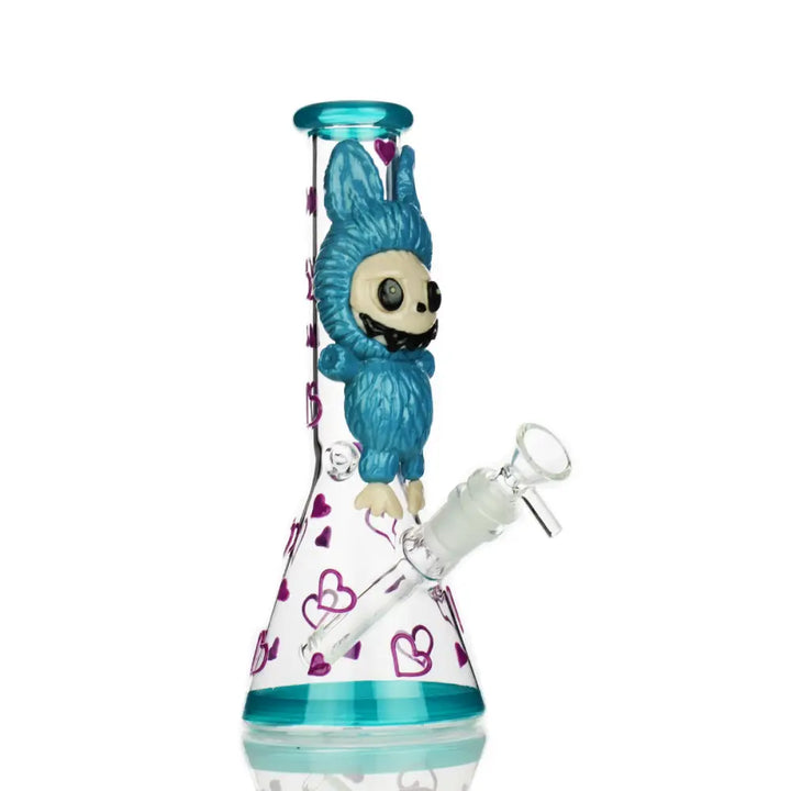 A full view of a clear glass beaker water pipe featuring a blue furry monster with a skull face and purple heart decals.