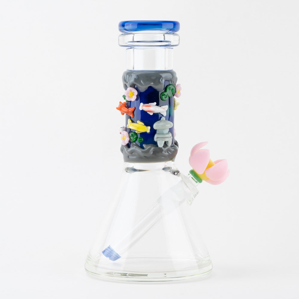 A clear glass beaker water pipe with a blue mouthpiece and a dark blue neck featuring a colorful koi pond design with fish, lotus flowers, and a pagoda. A pink lotus flower acts as the bowl handle.
