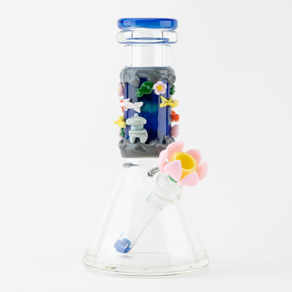 A clear glass beaker-style water pipe with a vibrant blue neck adorned with 3D elements depicting a koi pond scene, including colorful fish, a grey pagoda, and pink and yellow flowers. It features a pink lotus flower bowl.