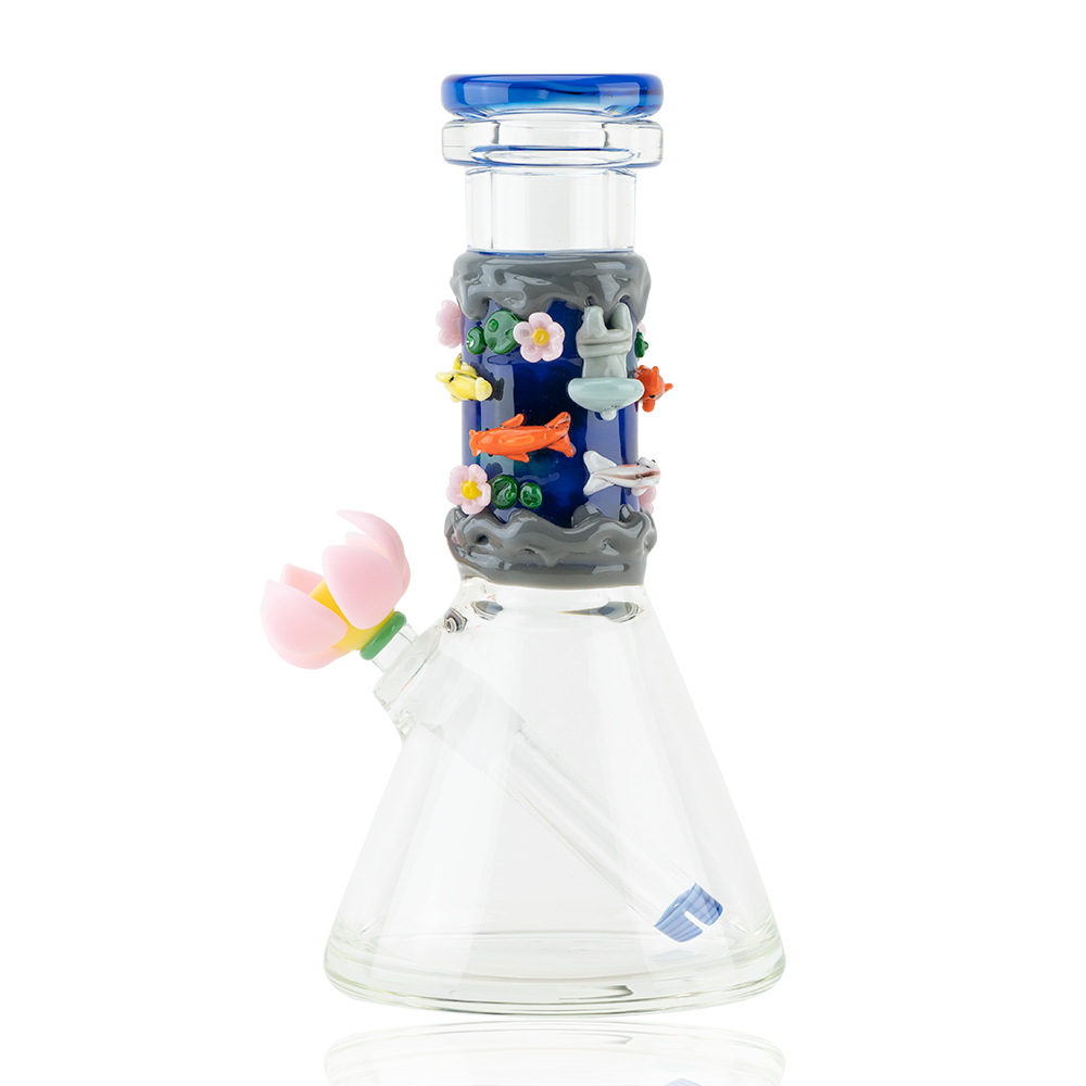 A clear glass beaker-shaped water pipe with a dark blue and grey section on the neck decorated with colorful glass koi fish, pink flowers, and underwater elements. It has a blue mouthpiece and a pink flower attachment on the side.