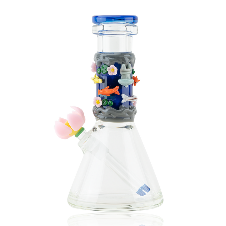 A clear glass beaker-shaped water pipe with a dark blue and grey section on the neck decorated with colorful glass koi fish, pink flowers, and underwater elements. It has a blue mouthpiece and a pink flower attachment on the side.