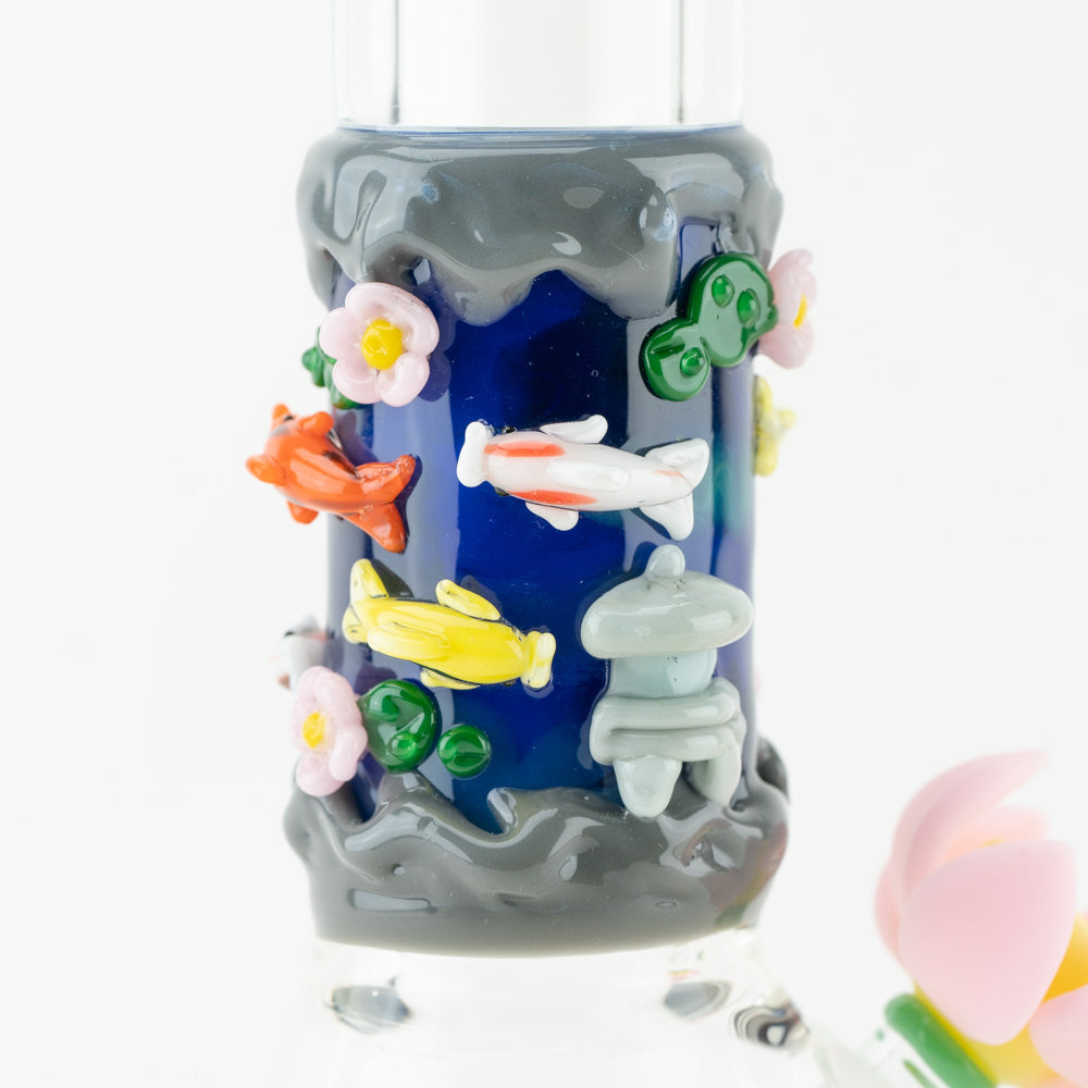 Detail shot of a dark blue glass water pipe section, adorned with colorful glass koi fish, green lily pads, pink and yellow flowers, and a grey stone lantern, resembling a koi pond.