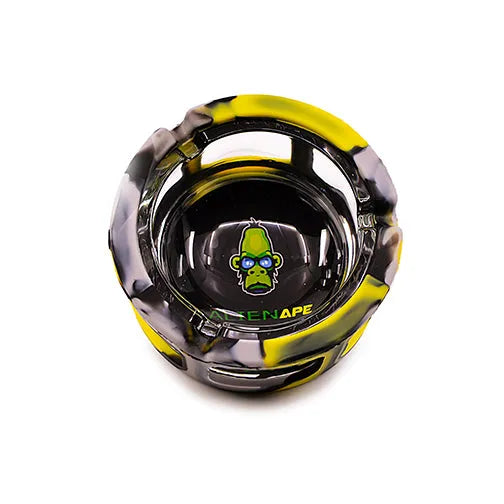 Top-down view of a round ashtray with a clear glass center and a yellow, black, and grey camouflage-patterned silicone sleeve. A green ape logo with blue glasses is visible in the center.