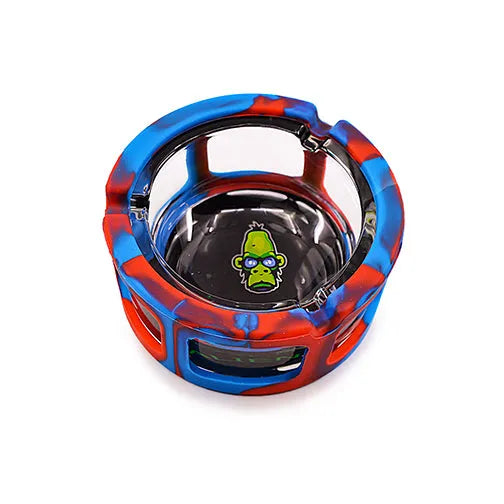 A round ashtray with a clear glass interior and a red and blue camouflage-patterned silicone sleeve. The bottom of the ashtray features a green cartoon monkey face wearing glasses.