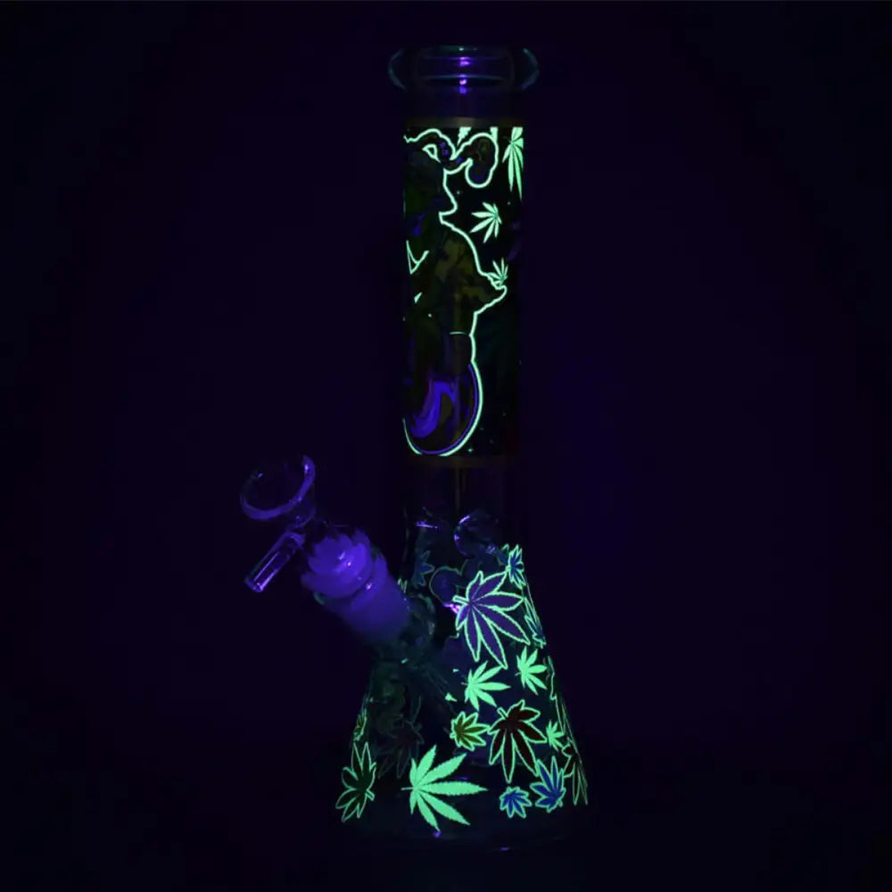 A glass beaker water pipe glowing in the dark, featuring bright green alien and cannabis leaf designs against a deep purple background.