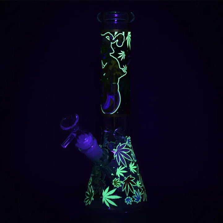 A glass beaker water pipe glowing in the dark, featuring bright green alien and cannabis leaf designs against a deep purple background.
