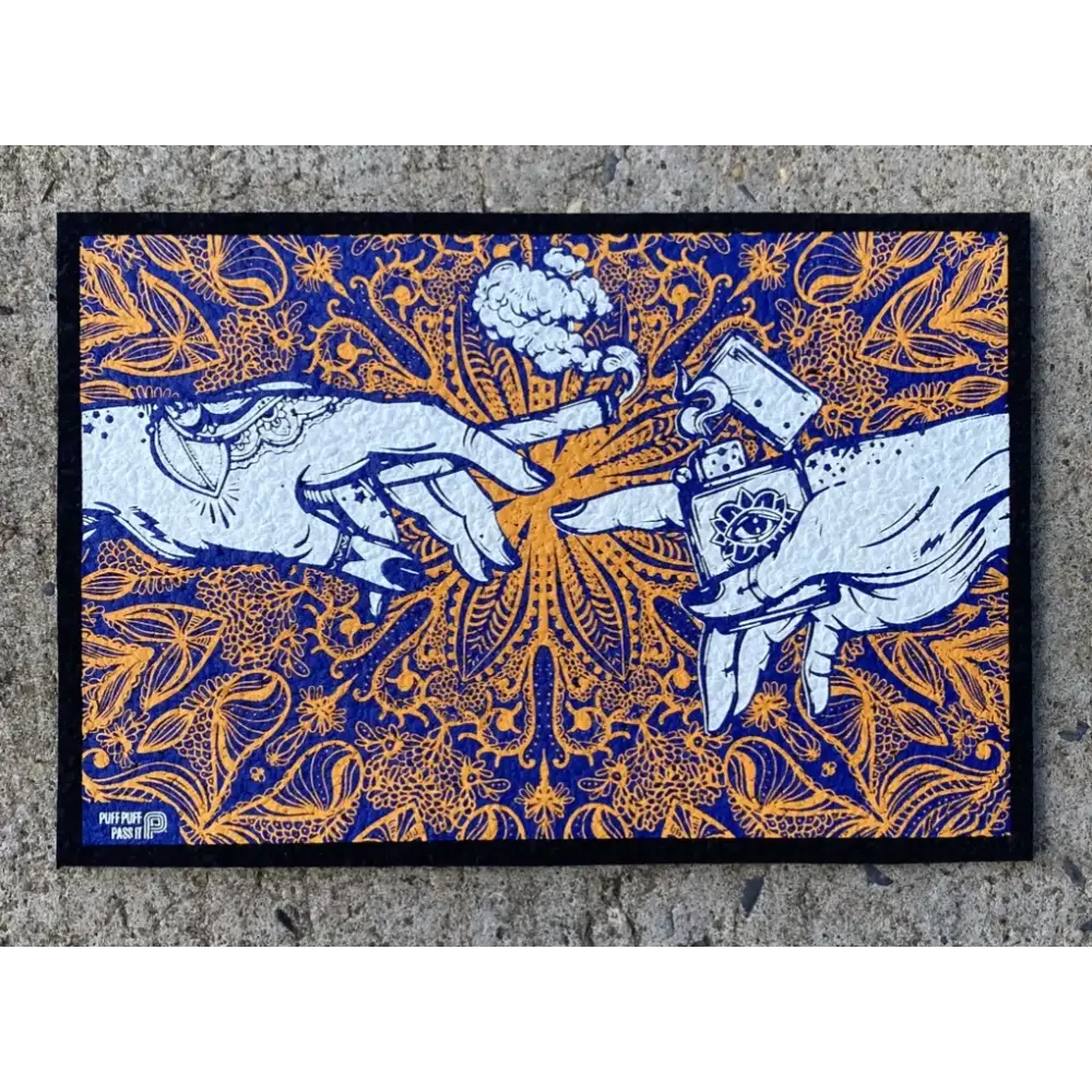 Rectangular mat featuring an illustration of two hands, one holding a lit joint and the other a lighter, in a 'Creation of Adam' style. The artwork is in white against a vibrant blue and orange mandala-like pattern with cannabis leaf motifs. The mat is placed on a concrete surface.
