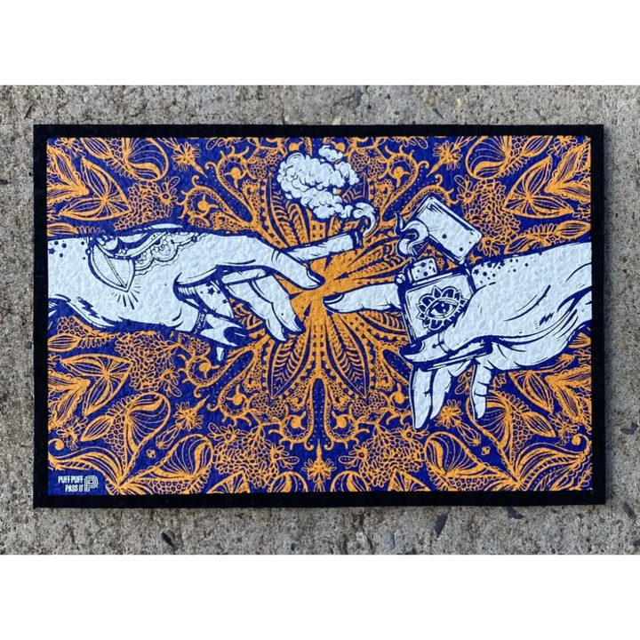 Rectangular mat featuring an illustration of two hands, one holding a lit joint and the other a lighter, in a 'Creation of Adam' style. The artwork is in white against a vibrant blue and orange mandala-like pattern with cannabis leaf motifs. The mat is placed on a concrete surface.