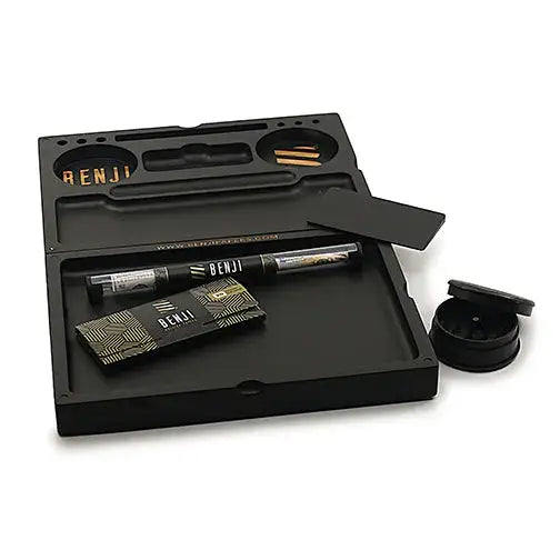 A black rectangular rolling tray kit is displayed open, revealing various compartments and accessories. Inside are a black tube with 'BENJI' text, a packet of rolling papers with a geometric pattern, and a small black grinder with its lid slightly ajar. The tray itself has 'RENJI' carved into one section.