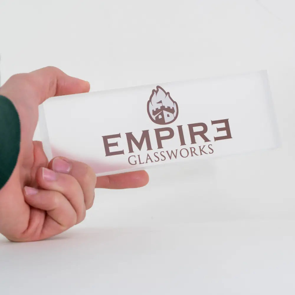 A hand holds a rectangular, frosted white acrylic plaque featuring a brown 'EMPIRE GLASSWORKS' logo with a castle icon.