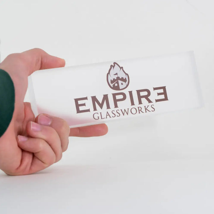 A hand holds a rectangular, frosted white acrylic plaque featuring a brown 'EMPIRE GLASSWORKS' logo with a castle icon.
