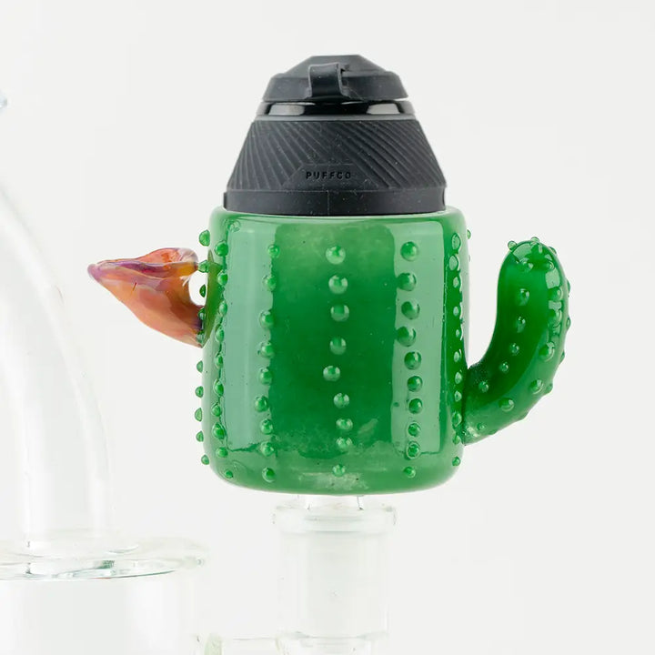 Close-up of a vibrant green glass attachment shaped like a cactus, featuring a small orange and purple flower, with a black Puffco vaporizer top attached.