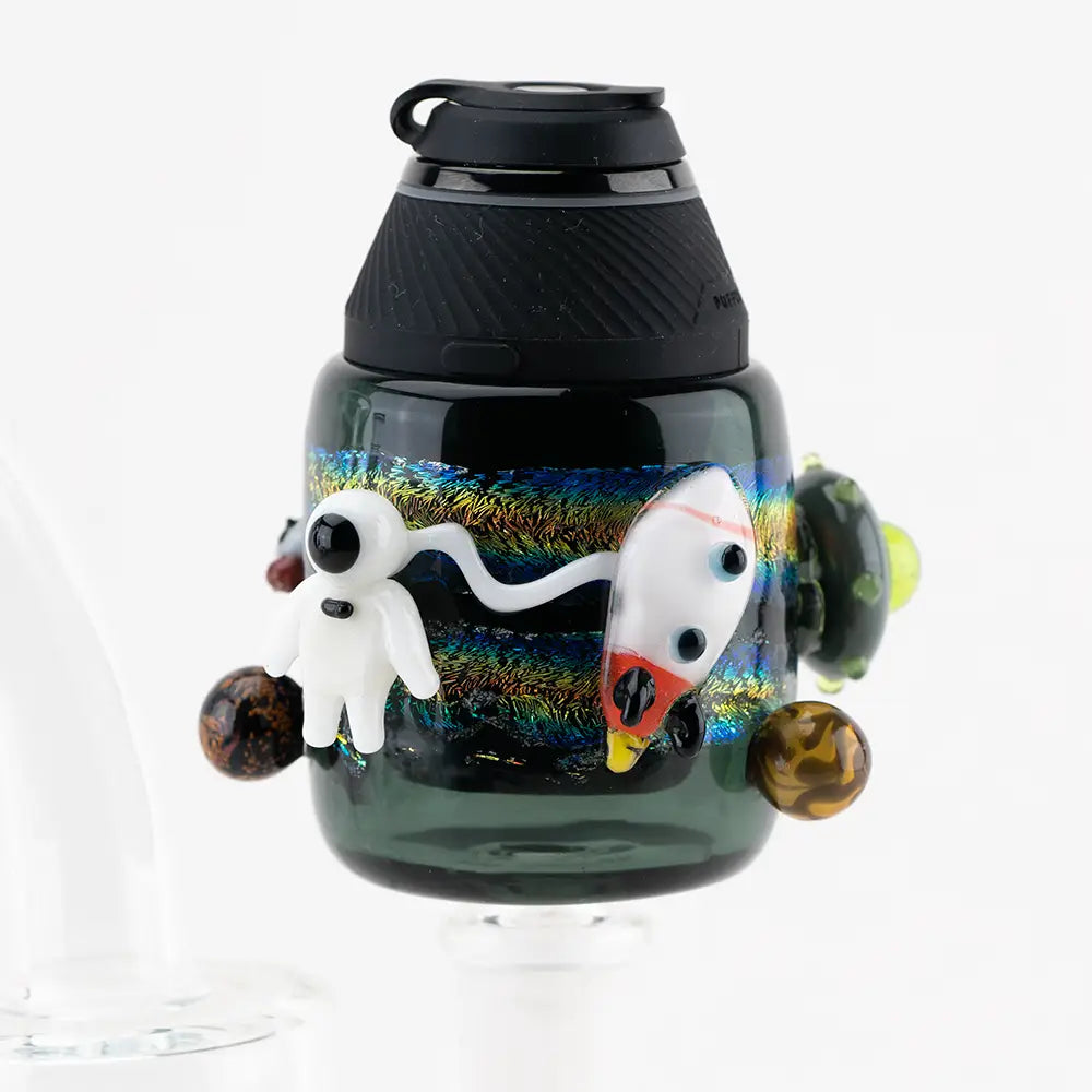 Close-up of a dark glass vaporizer attachment featuring a white astronaut, a white and red rocket, and brown planets against a shimmering, colorful galactic background.