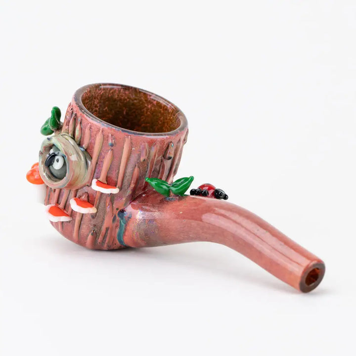 A whimsical glass hand pipe designed to resemble a tree stump, featuring an owl peeking from a hole, red-capped mushrooms, green leaves, and a ladybug on a white background.