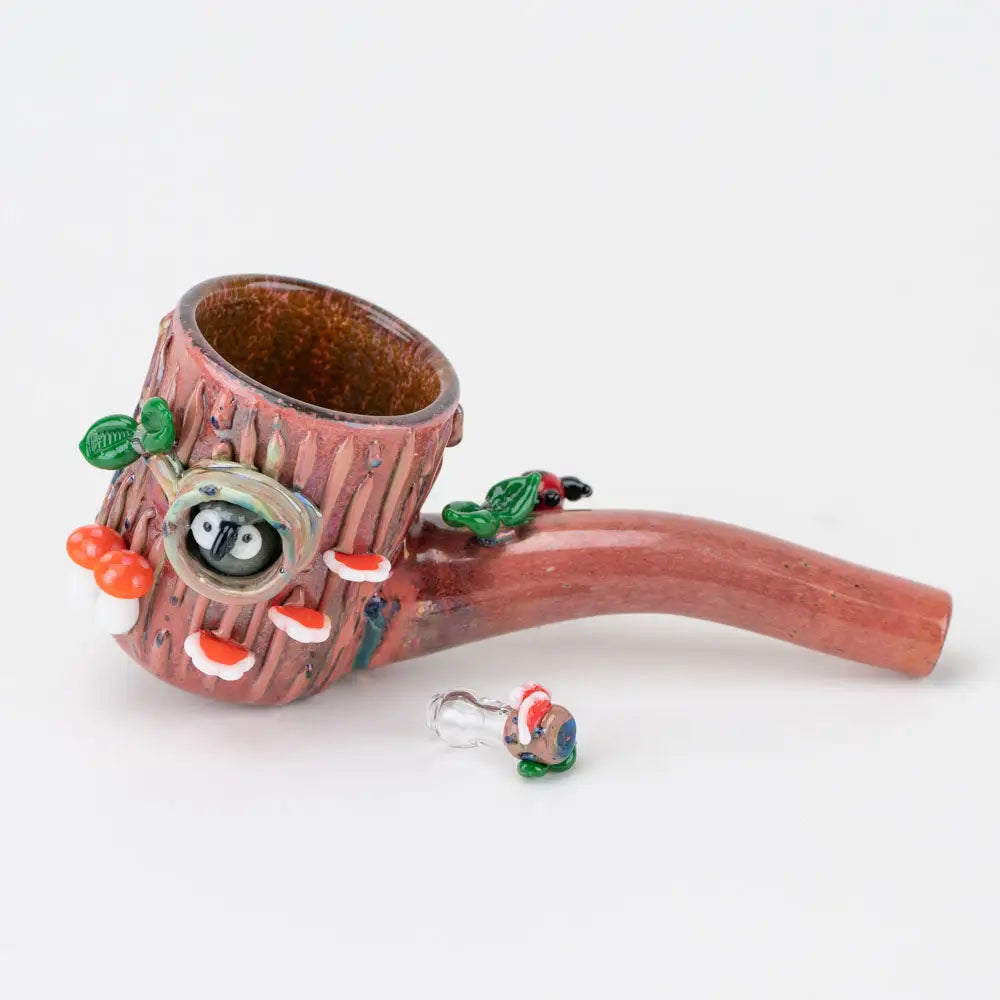 Close-up of a glass pipe shaped like a tree stump with an owl in a hollow, red and white mushrooms, and a green bird, with a small matching accessory piece in the foreground.