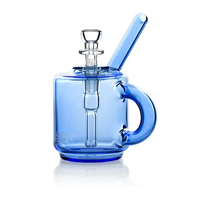 A vibrant blue glass bubbler, designed to resemble a coffee mug with a handle, stands upright on a reflective white surface, showcasing its clear glass downstem and bowl.