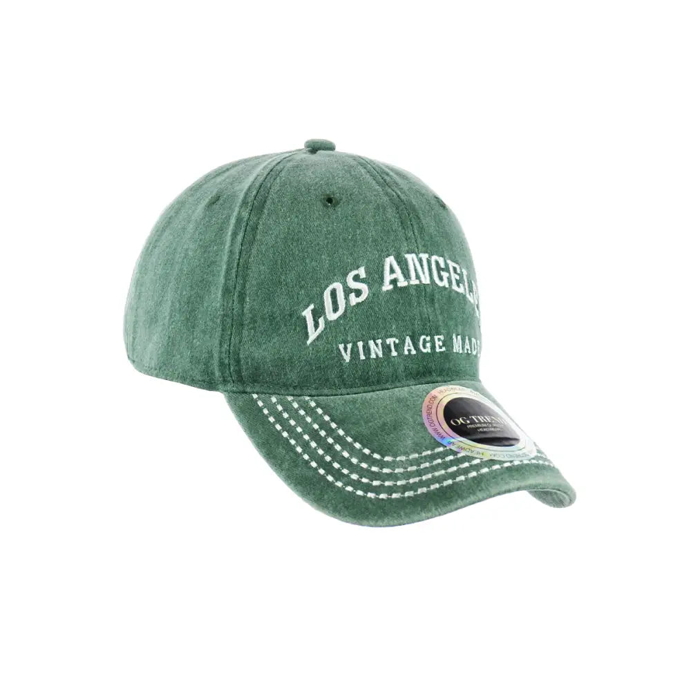 A close-up, studio shot of a distressed green baseball cap with 'LOS ANGELES' and 'VINTAGE MAD' embroidered in white on the front, featuring white stitching on the brim and a holographic tag.