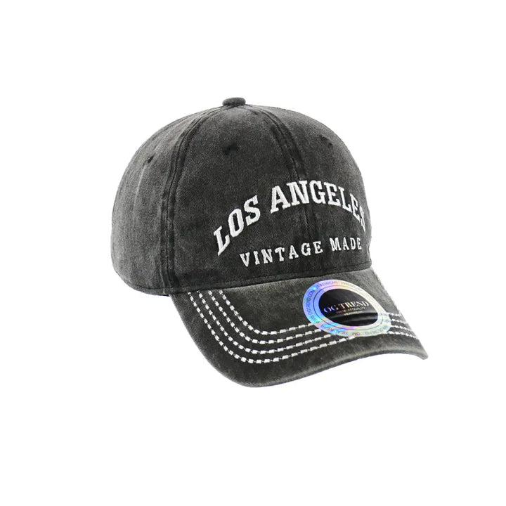 A dark gray, distressed baseball cap with 'LOS ANGELES' and 'VINTAGE MADE' embroidered in white on the front, featuring white stitching on the brim and a holographic sticker.