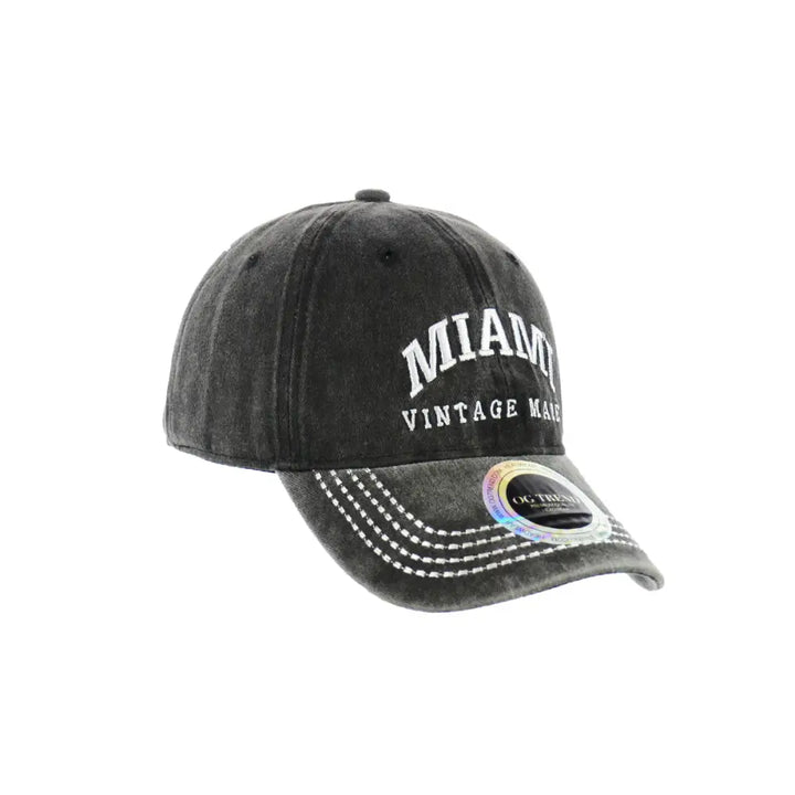 A dark grey, distressed-look baseball cap with 'MIAMI' in large white arched letters and 'VINTAGE MADE' in smaller white letters embroidered on the front. The brim features multiple rows of white stitching, and a holographic sticker is visible on the right side of the brim.
