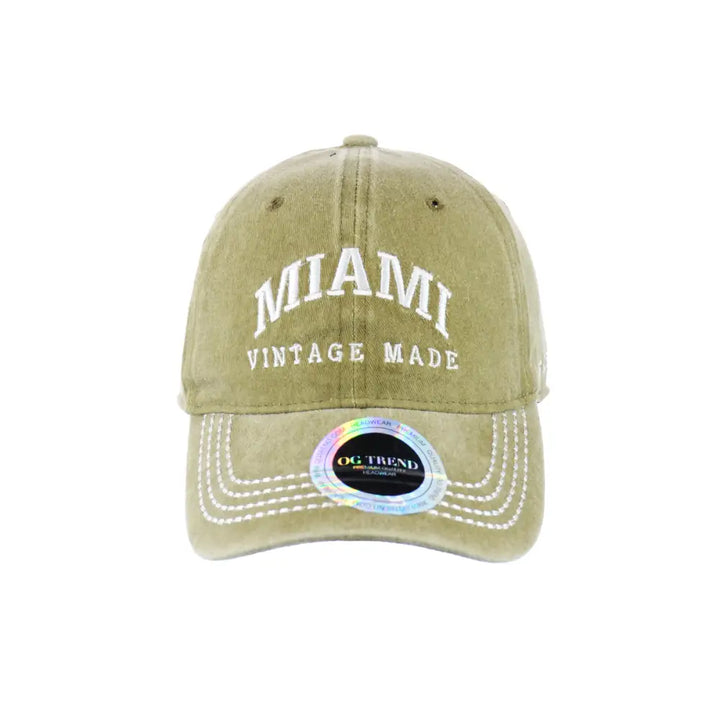 Front view of a distressed olive green baseball cap with 'MIAMI VINTAGE MADE' embroidered in white, featuring visible stitching on the brim and a holographic 'OG TREND' sticker.