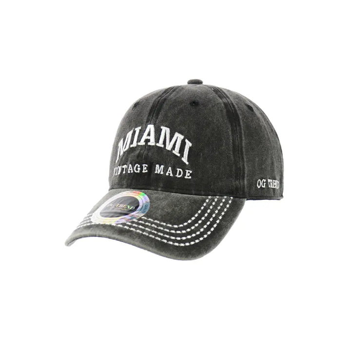 Close-up product image of a dark charcoal grey baseball cap with 'MIAMI' embroidered in white on the front, and 'VINTAGE MADE' below it. The brim has white stitching and a holographic sticker.