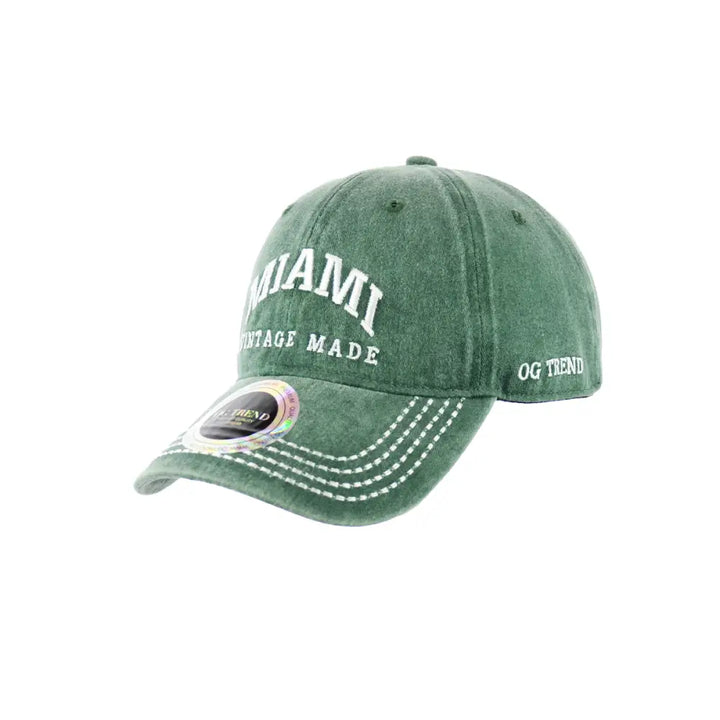 A dark green, distressed-look baseball cap with 'MIAMI' and 'VINTAGE MADE' embroidered in white on the front, and 'OG TREND' on the side. The brim has white stitching and a holographic sticker.