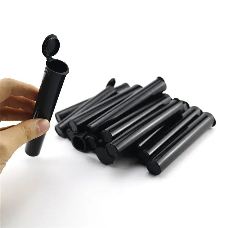 A hand holds a single black plastic pre-roll tube with its hinged lid open, while a pile of many similar black tubes rests in the background.