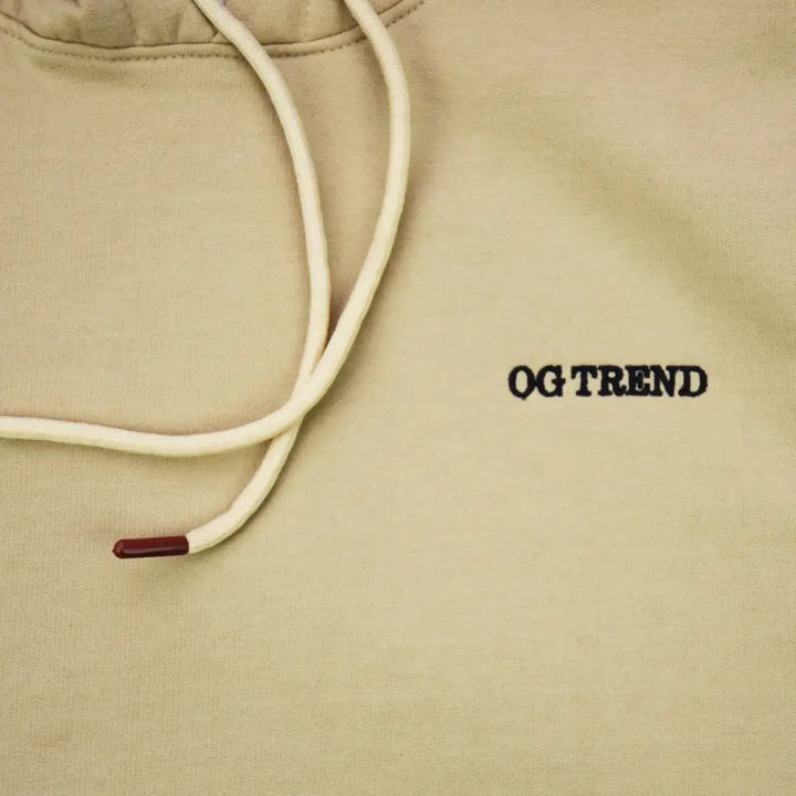 Close-up detail shot of a light tan hoodie, showing the cream-colored drawstrings with dark red aglets and the embroidered black text 'OG TREND' on the chest.
