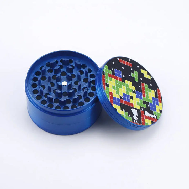 An open blue metal herb grinder with a pixelated, 8-bit style design on its lid, featuring colorful geometric shapes and a small figure.
