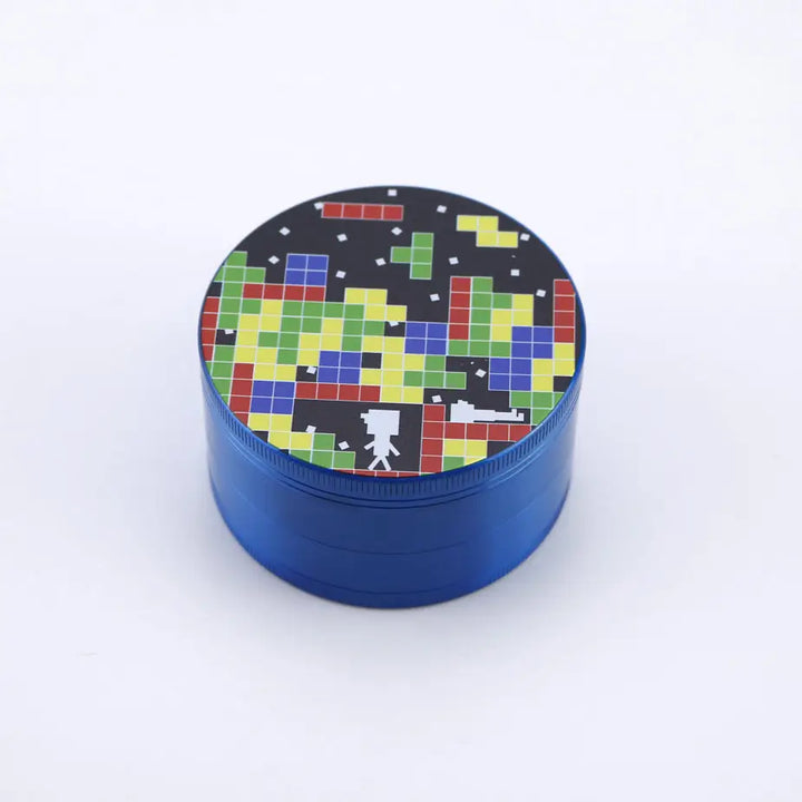 Close-up view of a blue metal herb grinder, showing the top lid with a colorful pixelated design resembling Tetris blocks and a small white pixelated figure.